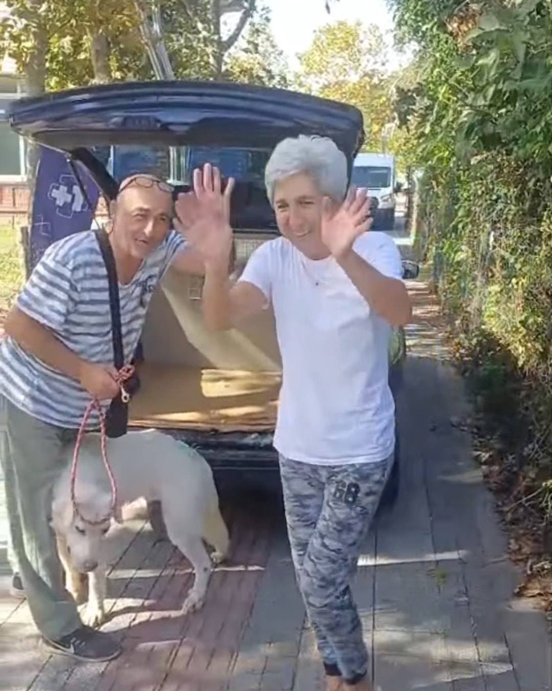 Gülümseyerek el sallayan iki kişi ve yanlarında iki köpek, arkada bagajı açık araç.