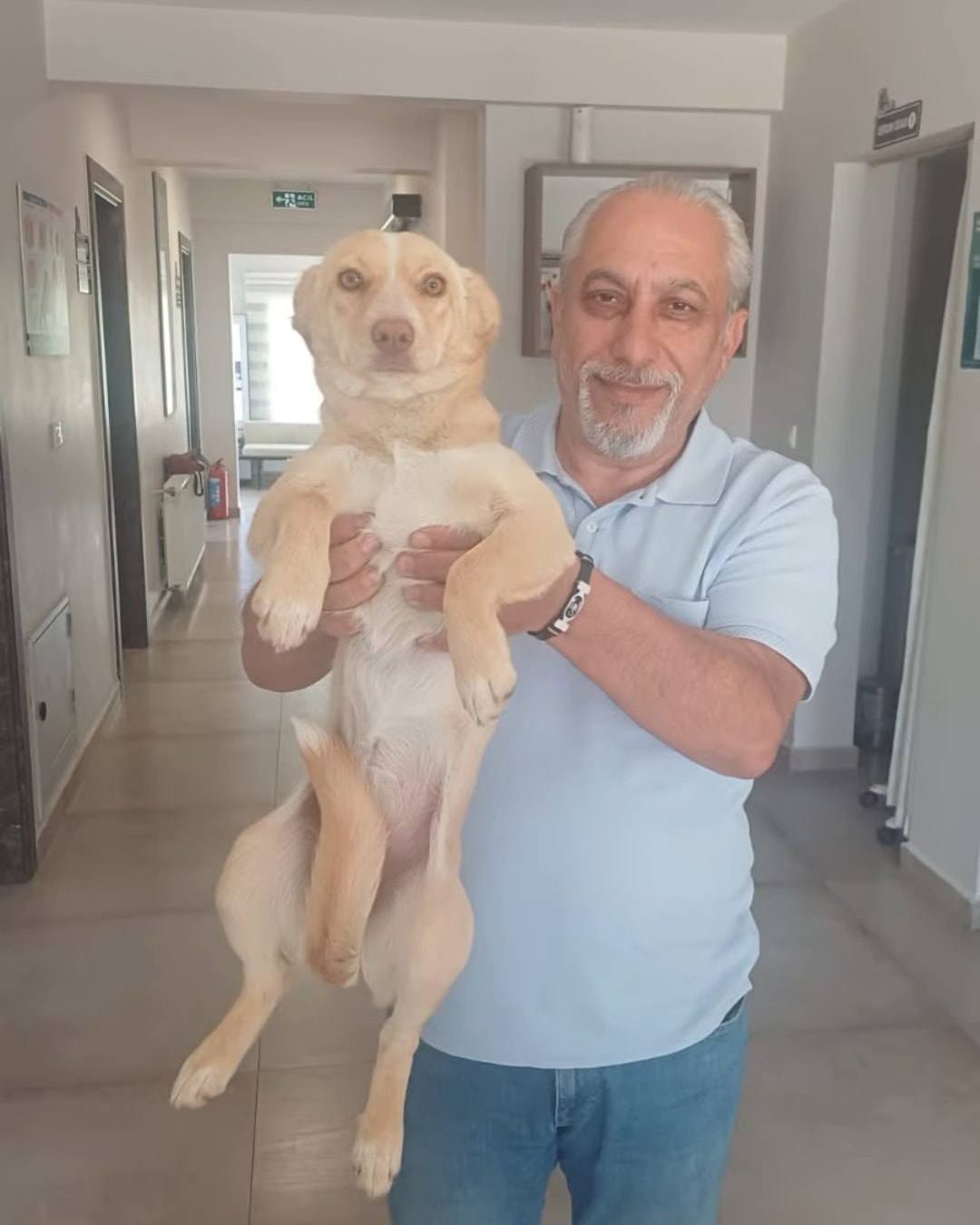 Gülümseyen bir adam kucağında sarı tüylü bir köpeği kameraya gösteriyor.